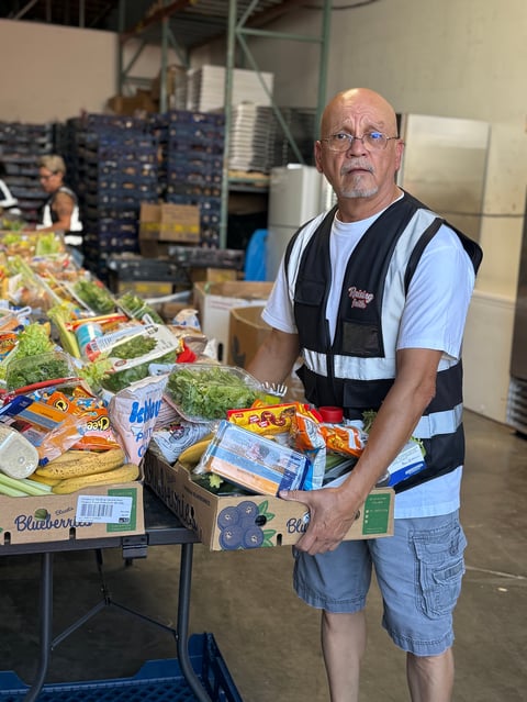 Warehouse worker in safety vest standing at table with packaged produce and food items in industrial setting