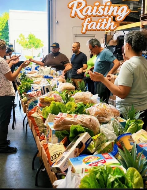 Community members gather around a table of fresh produce and groceries at a Raising Faith food distribution event in a warehouse setting