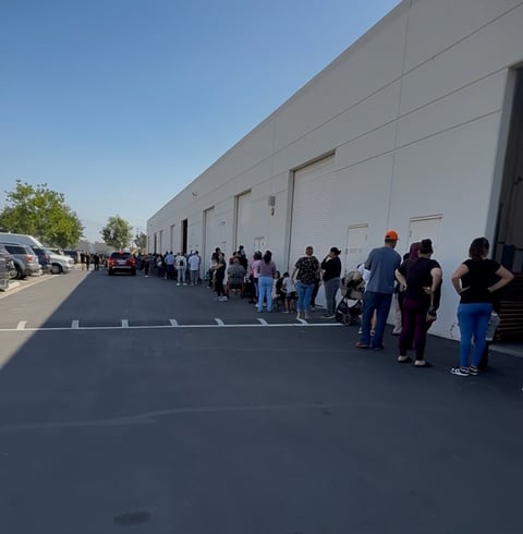 Line of people queuing outside a large industrial warehouse building on a sunny day