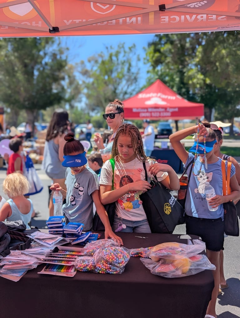 Children at outdoor community event receiving prizes and merchandise at a vendor booth under red tent with trees and park setting in background