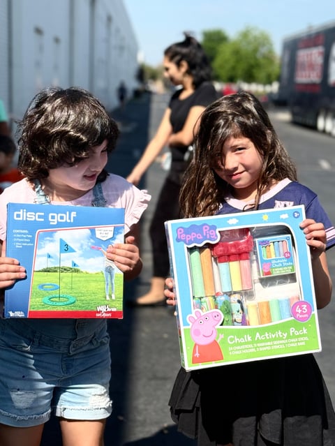 Two young children holding toy boxes at an outdoor event, with vehicles and adults visible in the background