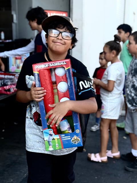 Child wearing glasses holding a pop-up baseball game toy while other children stand in background at an indoor event